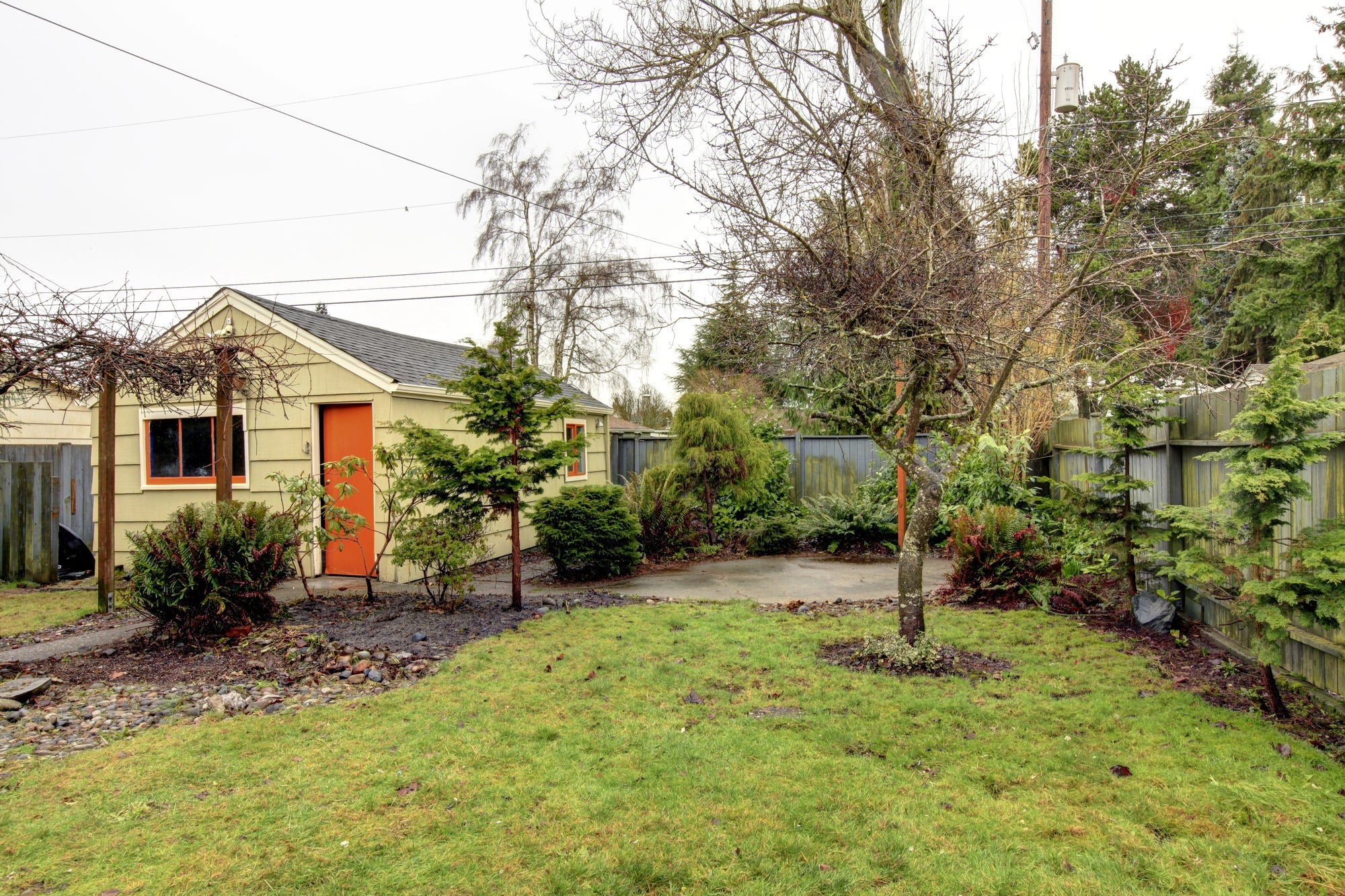 Blick auf einen leicht verwilderten Garten mit Rasenfläche, Sträuchern und Bäumen, im Hintergrund ein kleines Gartenhaus mit orangefarbener Tür.